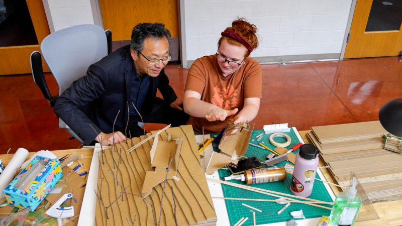 Dean Liu and a student looking at cardboard model on table together.