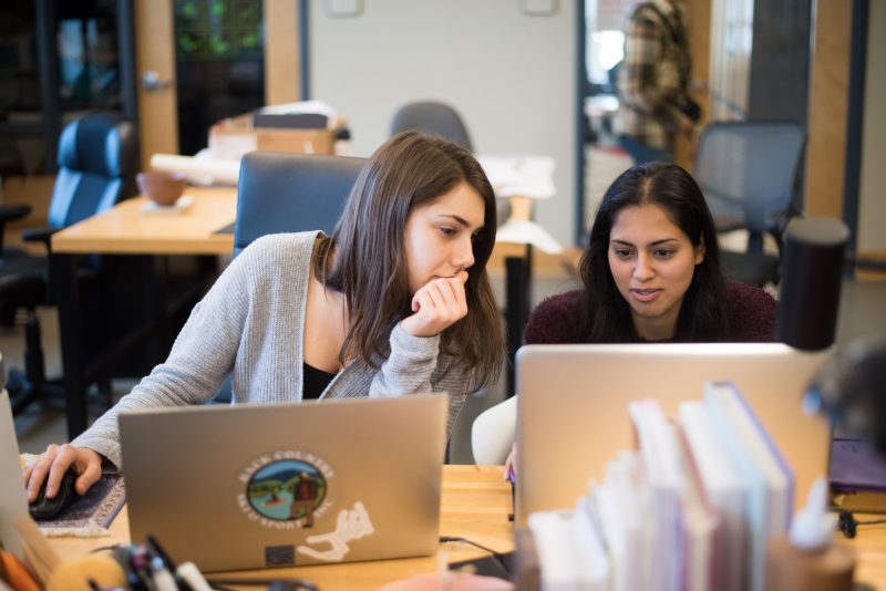 Two interior design students sitting at a table with laptops, one engaging with the other, both looking at one laptop screen