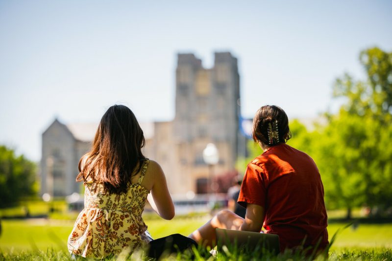 The backs of two students sitting on the drillfield looking toward Burruss Hall.