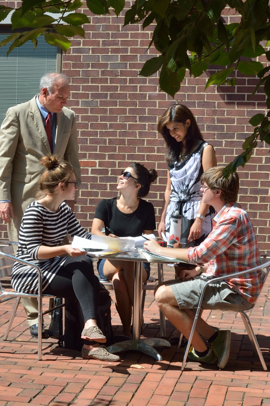An instructor oversees four students comparing notes at an outdoor dining table.
