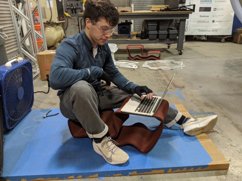 A student in a workshop sits with their laptop atop a metallic frame.
