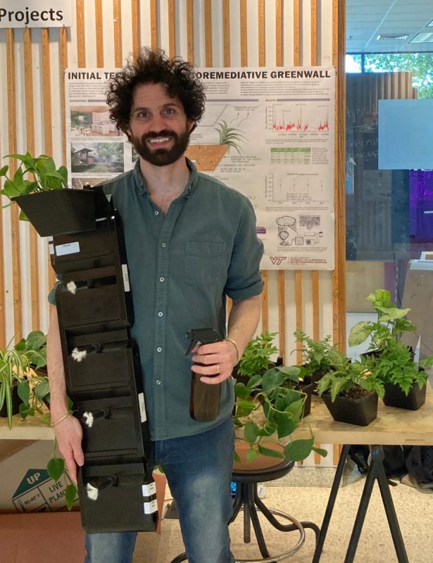 Zachary Gould holding a stack of plants with a table of plants behind him and a screen showing an animation of plants + research.