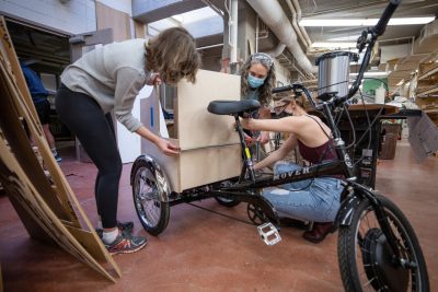 Three people make adjustments to a bike with a box on the back of it.