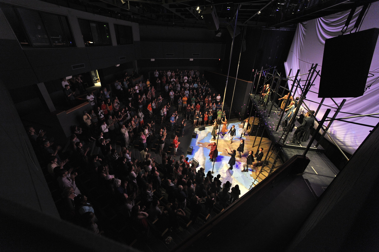 Full view of stage with performers, rafters with crew, and audience around the base of the stage.
