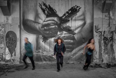 Three people run toward the camera in front of a large black-and-white mural of two clenched hands holding broken structures that resemble city buildings. The person in the center is sharply focused, wearing dark clothes with teal suspenders, while the figures on either side are blurred in motion. 