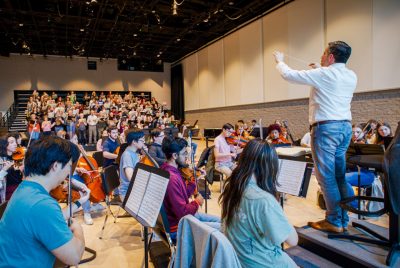 Musicians and singers rehearsal indoors watching their conductor on the podium.