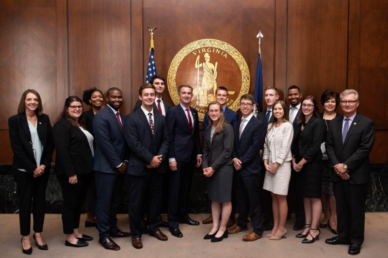 Several young professionals pose with Virgiia Governor Ralph Northam and other dignitaries in front of the seal of the Commonwealth of Virginia in a conference room in Richmond, Virginia