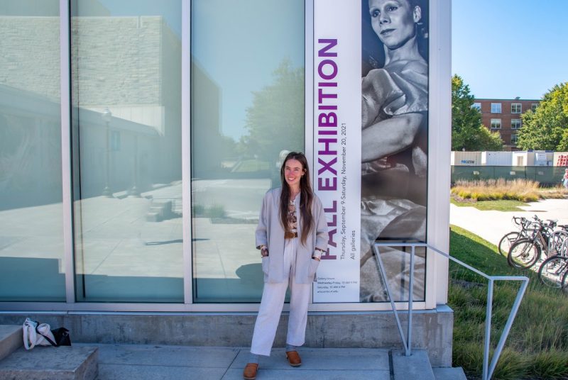 Student Ali Palin wears a light colored blazer and pants and stands outside of a section of windows, the last pane featuring a panel with a black and white photo that says fall exhibition.