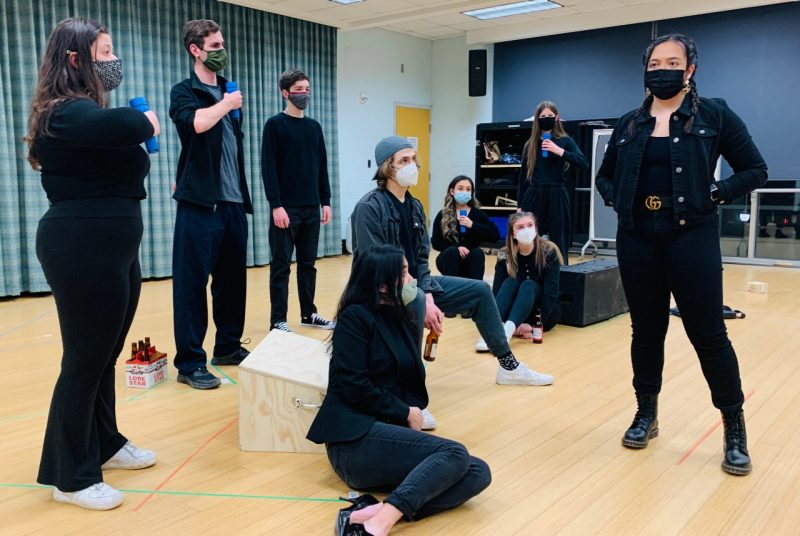 Nine college students, all dressed in black and wearing masks, some sitting and some standing, rehearse a scene from a play in a room with wooden floor and blue walls
