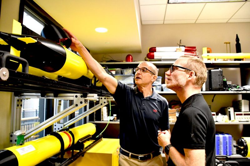 Two men stand before a rack of multiple, yellow, robotic submarines inside a Torgersen Hall lab. One of the men, in the background, points out features on the submersible to the other.