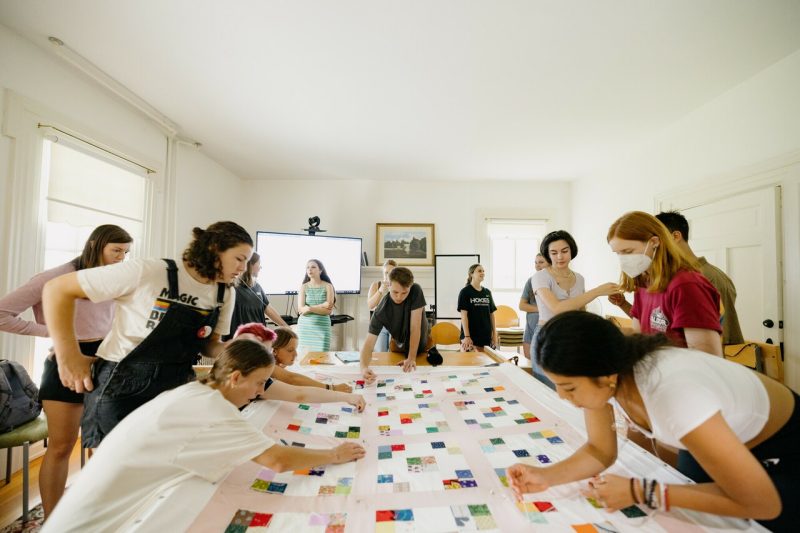 More than a dozen students gather around a quilt tacked to a quilting frame as they tie it.