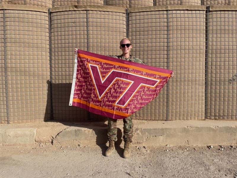 U.S. Army 1st Lt. Meredith Oakes Moughan smiles while holding a Virginia Tech flag. She is wearing her camouflage uniform and sunglasses.