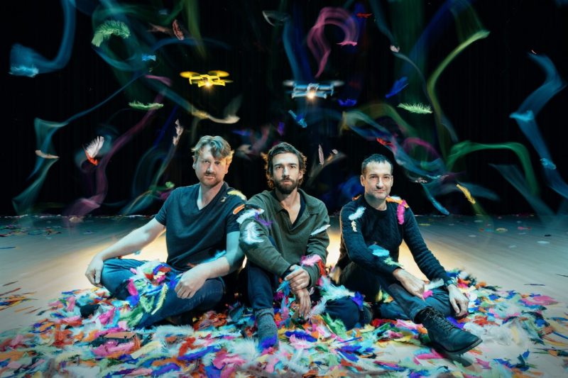 Three faculty members who collaborated on Daedalus Dreams—Zach Duer, Scotty Hardwig, and Eric Handman—smile at the camera while yellow and blue drones hover above their heads and brightly colored feathers fall 