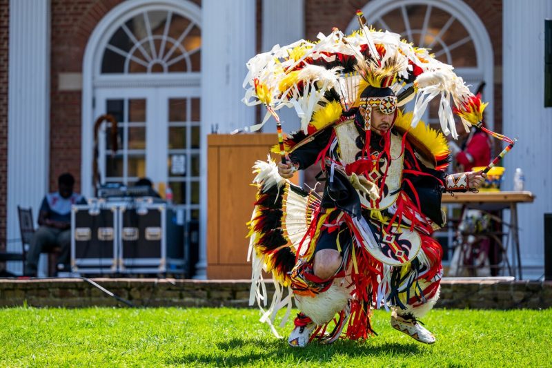A male dancer dancing in full red, black, yellow, and white regalia on a green lawn in front of a brick building. 